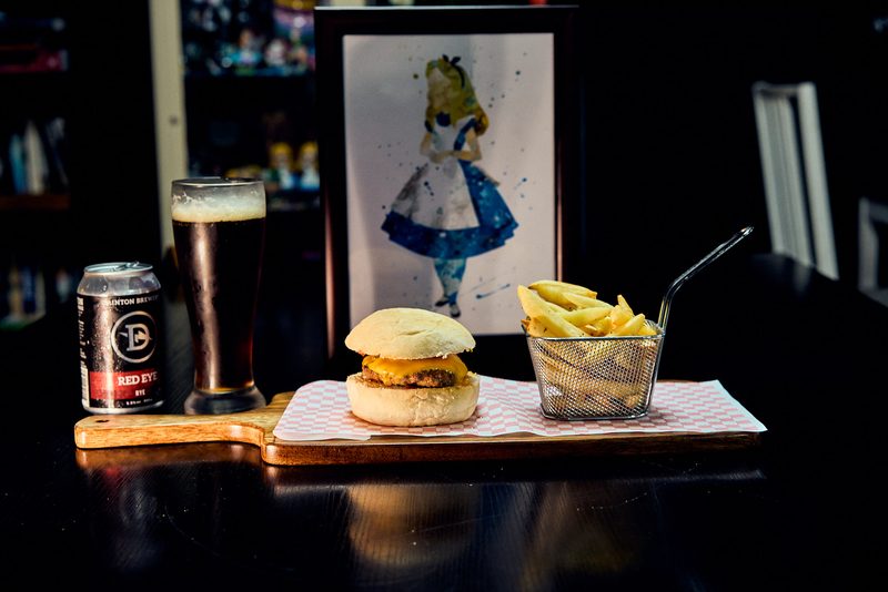 A burger on a serving board with a basket of fries, a can and a dark beer, an illustrated print behind