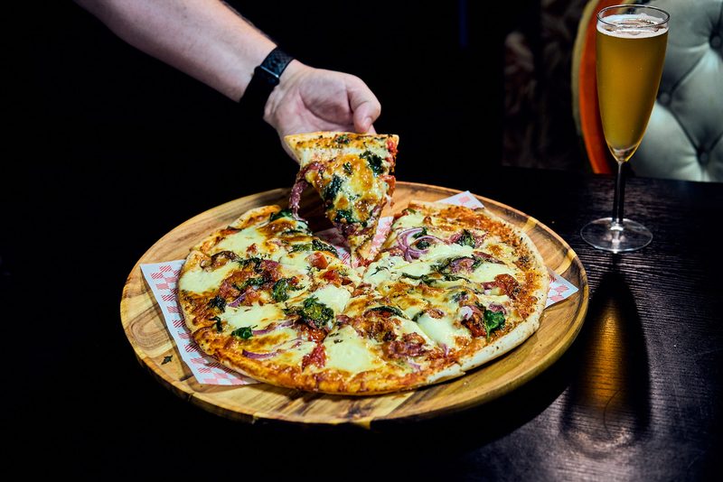 A hand lifting a slice from a pizza on a wooden board, a glass of beer beside it on a dark table